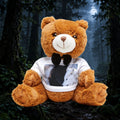 A large brown plush teddy bear sits in a dark forest scene, wearing a white T-shirt with a black bow tie and a printed photo of a black and white dog on the chest.