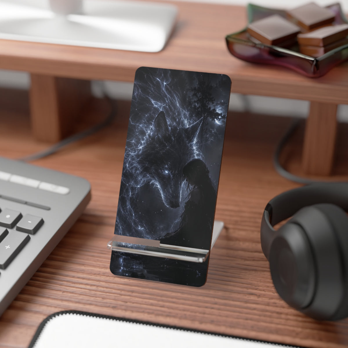 A black phone stand with a dark cosmic wolf design, shown holding a smartphone on a wooden desk with a keyboard, headphones, and other desk accessories in the background.